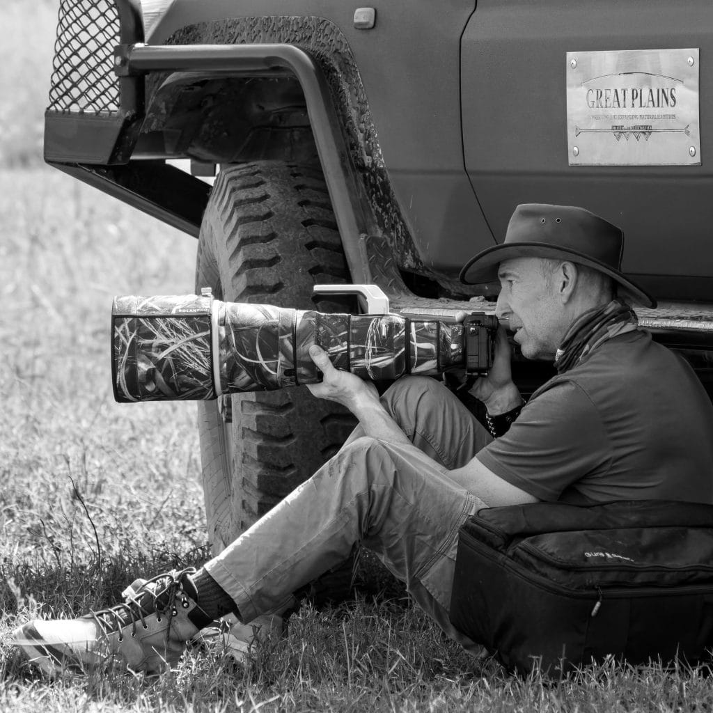A Black & White photograph of the author sitting in front of a safari vehicle with a massive zoom lens in his hands.